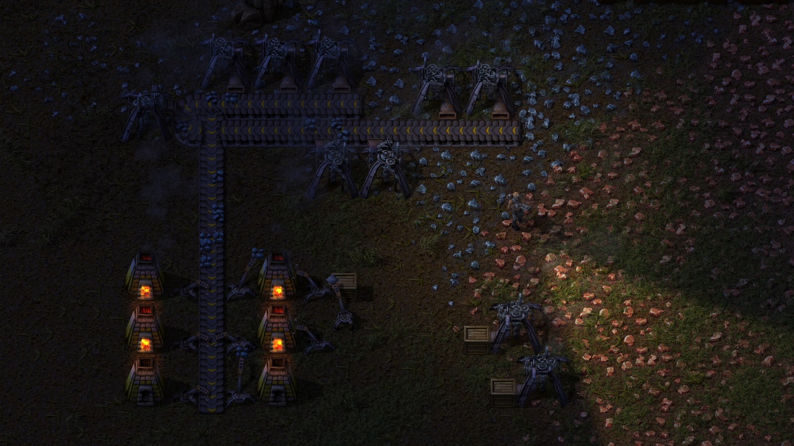 An early Factorio base at night: two columns of three stone furnaces glowing orange, fed by mechanical arms from a yellow transport belt running up the middle of the screen, with burner mining drills on patches of blue-grey ore at the top and orange-red ore at the right, wooden crates beside them, and the player character lit by a small lamp in the dark grass