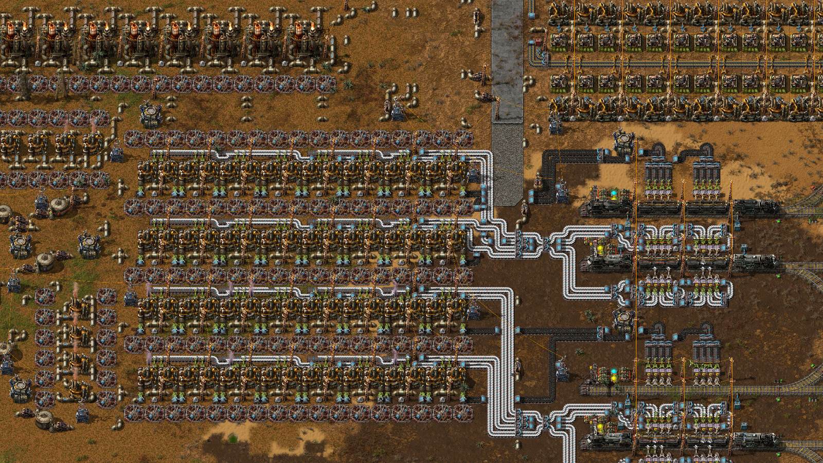 A large Factorio factory by day: several long rows of assembling machines and chemical plants linked by mechanical arms, white and blue pipe runs curving between them, overhead power poles and wires, and trains standing at stations on rail lines to the right, all built on brown dirt and patches of grass