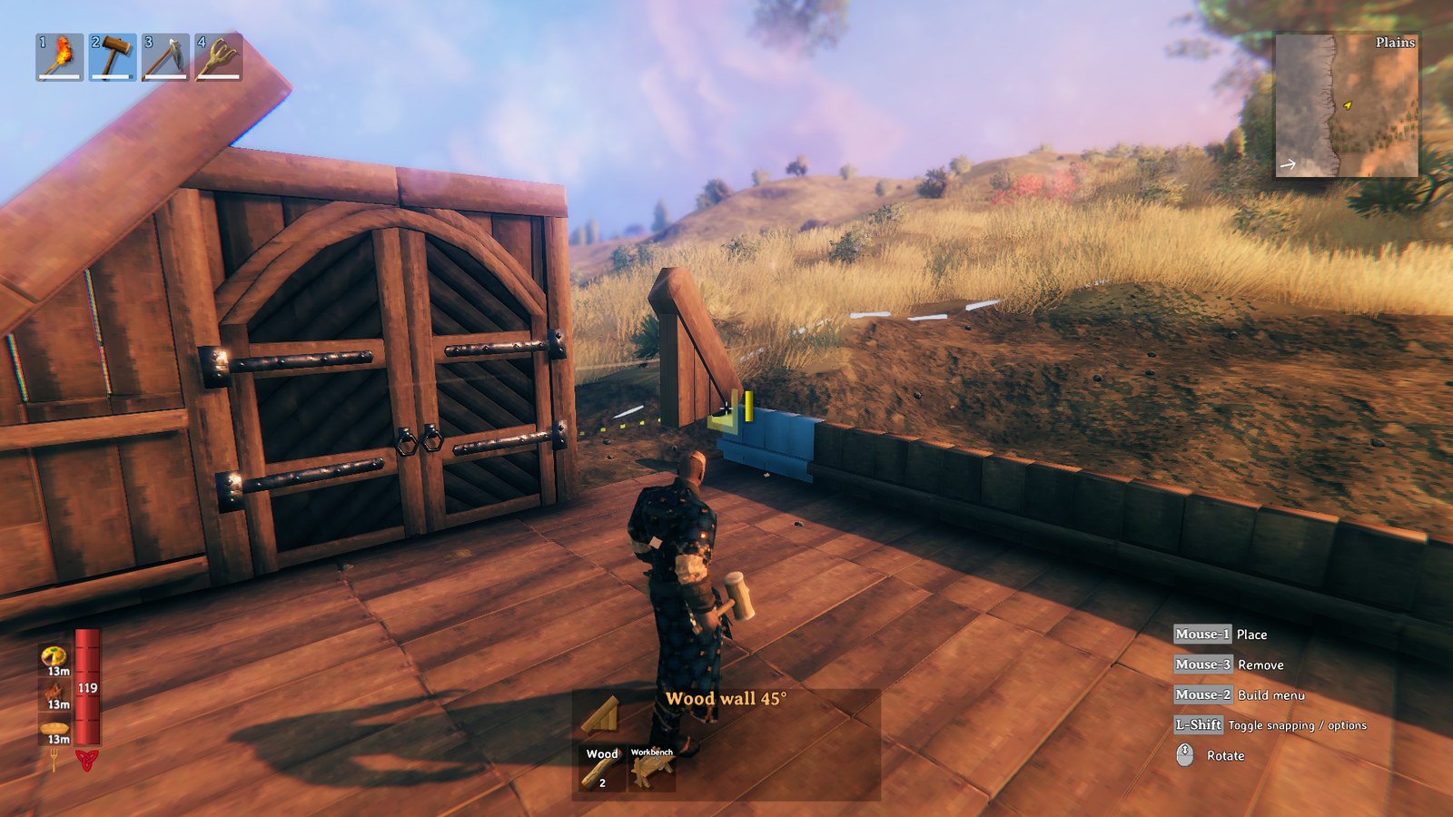 A Valheim player in build mode on a wooden floor, placing a wood wall at 45 degrees: the translucent blue placement ghost sits beside a large arched double door, a stone wall runs along the edge of a plains biome of golden grass, the hotbar shows a torch, hammer, pickaxe and cultivator, and the control hints read Mouse-1 Place, Mouse-3 Remove, Mouse-2 Build menu, L-Shift Toggle snapping